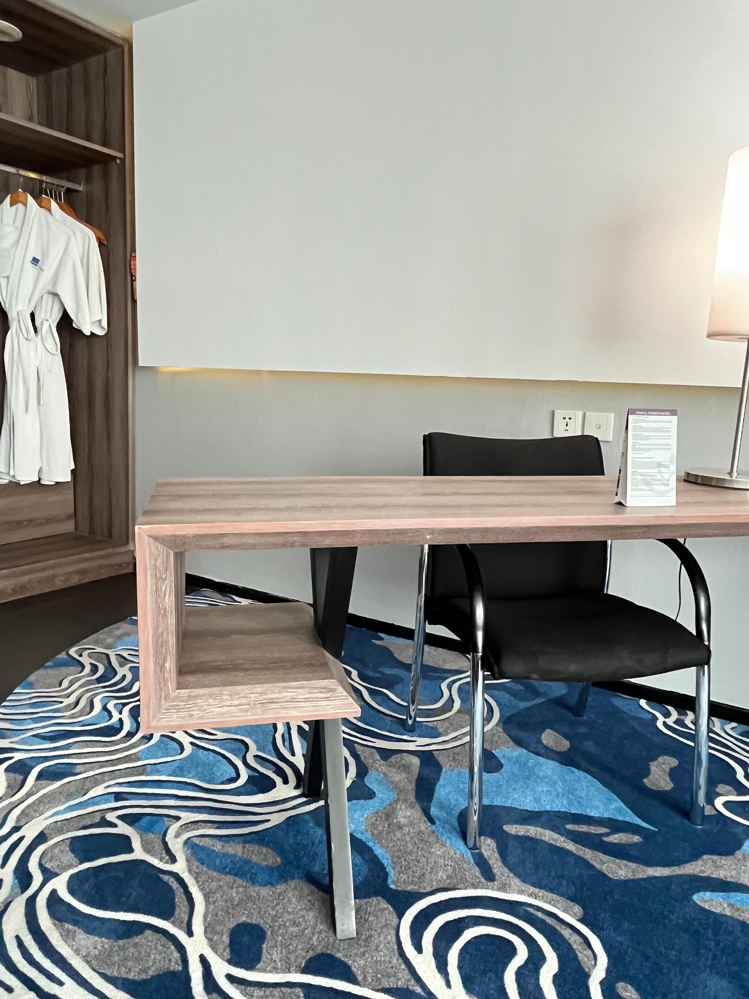 A hotel room with a wooden table, black chair, and a blue patterned carpet.