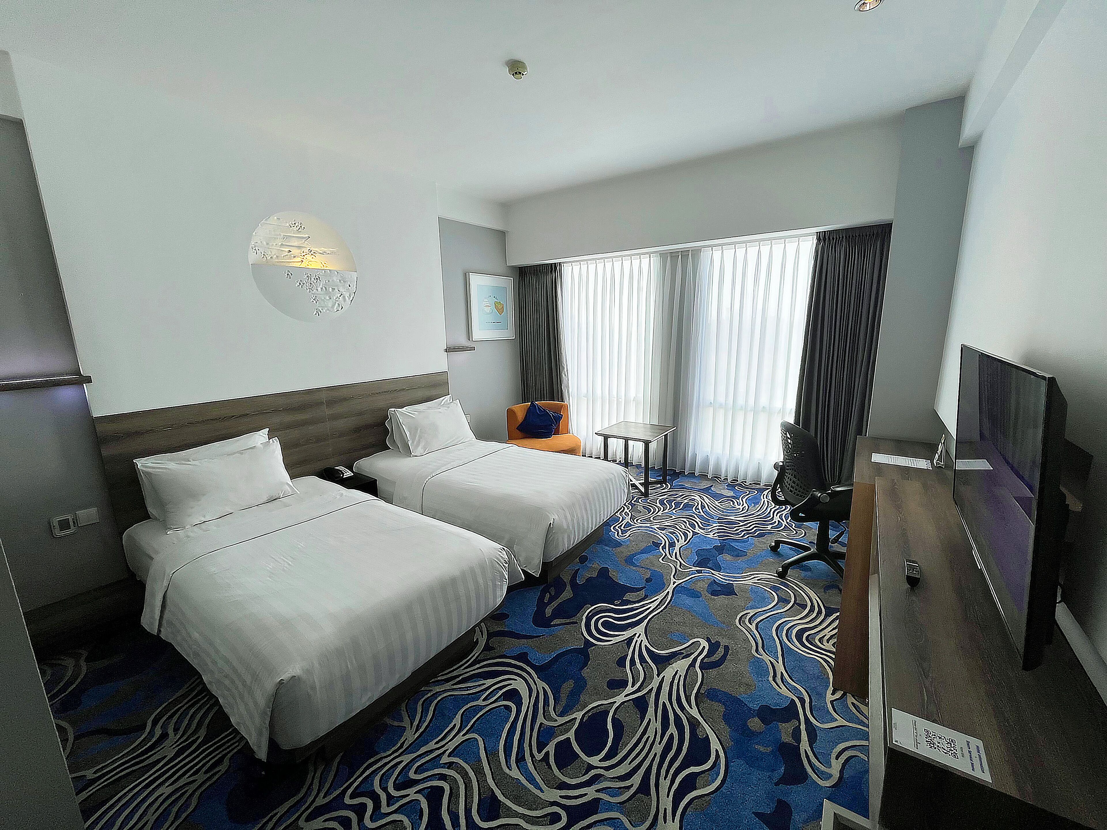 Quarto de hotel com duas camas, uma escrivaninha, uma cadeira, televisão e uma janela grande com cortinas.