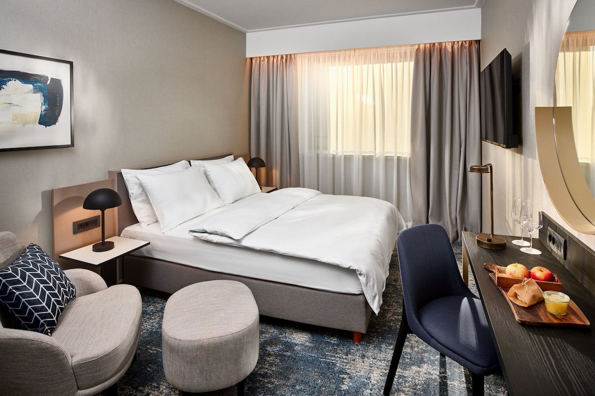 A modern hotel room with a large bed, a desk with a chair, a round ottoman, and a dining area with fruits and a glass of juice.