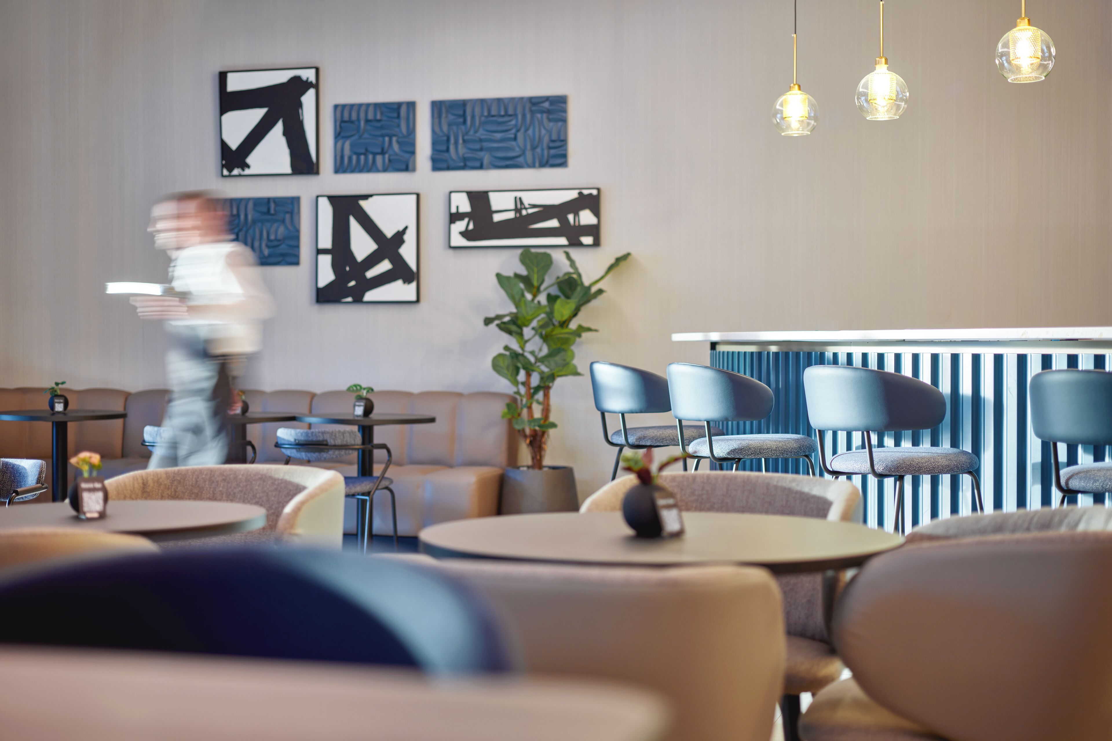 Un restaurante moderno con mesas redondas y sillas azules, una zona de bar y arte abstracto en las paredes.