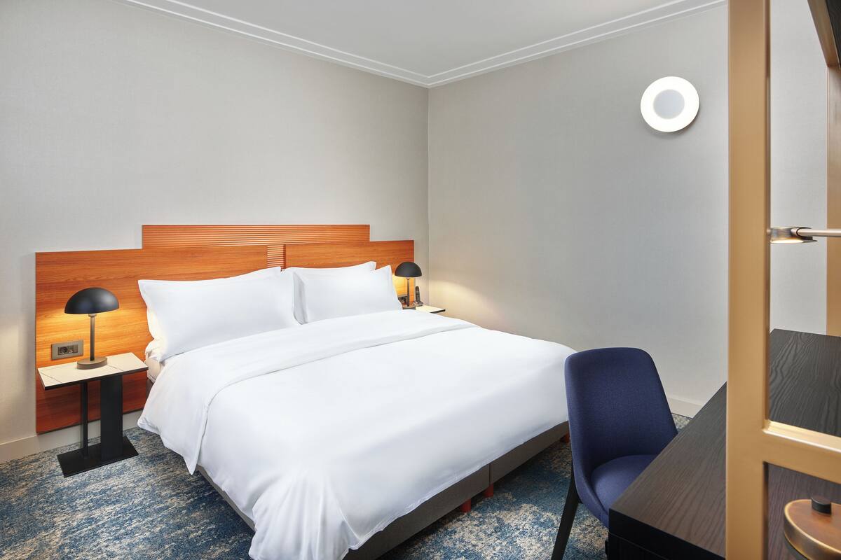 A hotel room with a wooden headboard bed, white bedding, a blue chair, and a small table with a lamp.