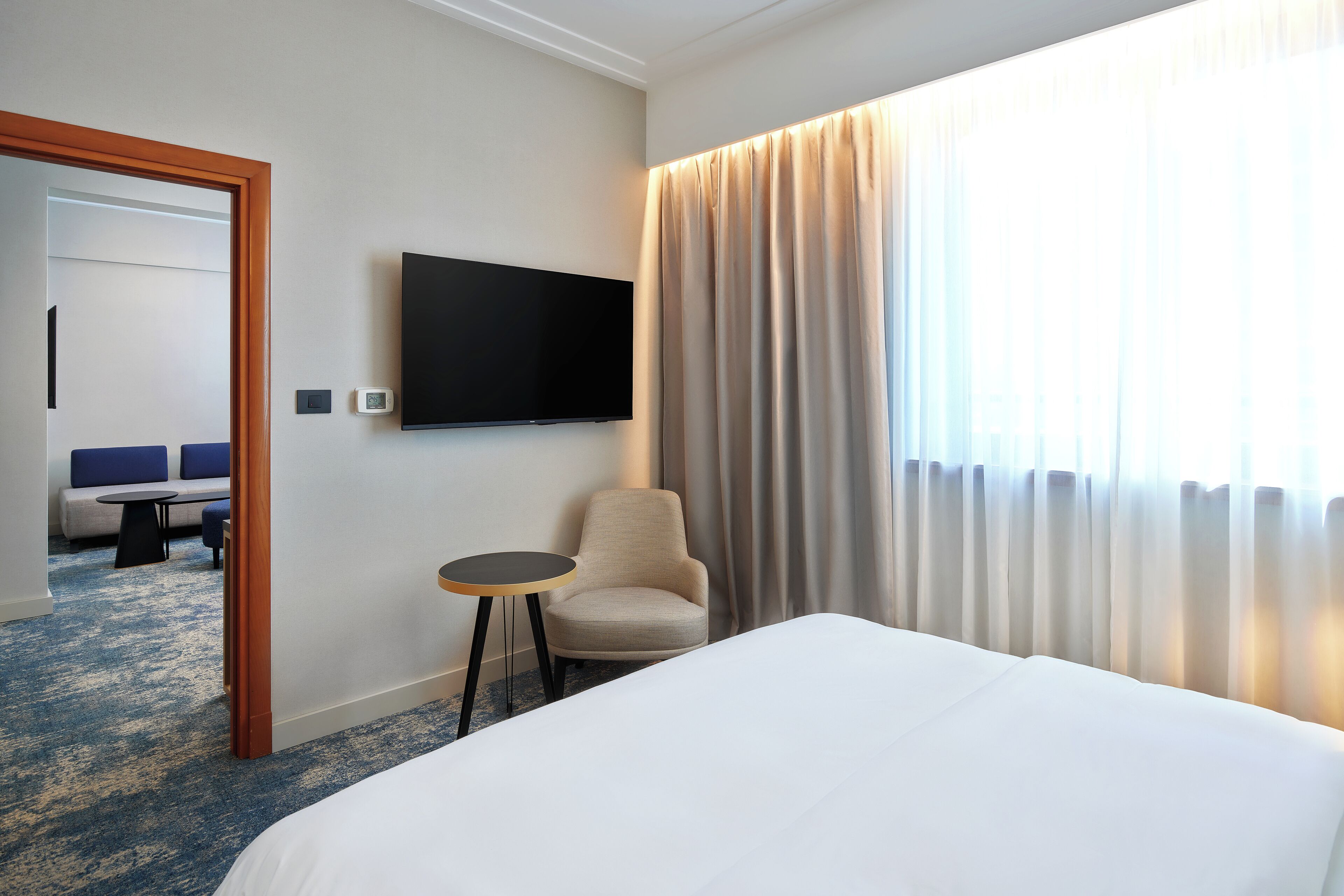 A hotel room with a bed, a chair, a small table, a TV mounted on the wall, and a view of a balcony with curtains.