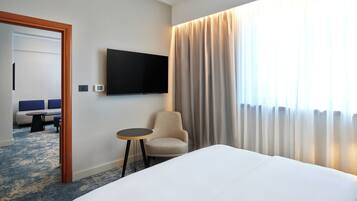 A hotel room with a bed, a chair, a small table, a TV mounted on the wall, and a view of a balcony with curtains.