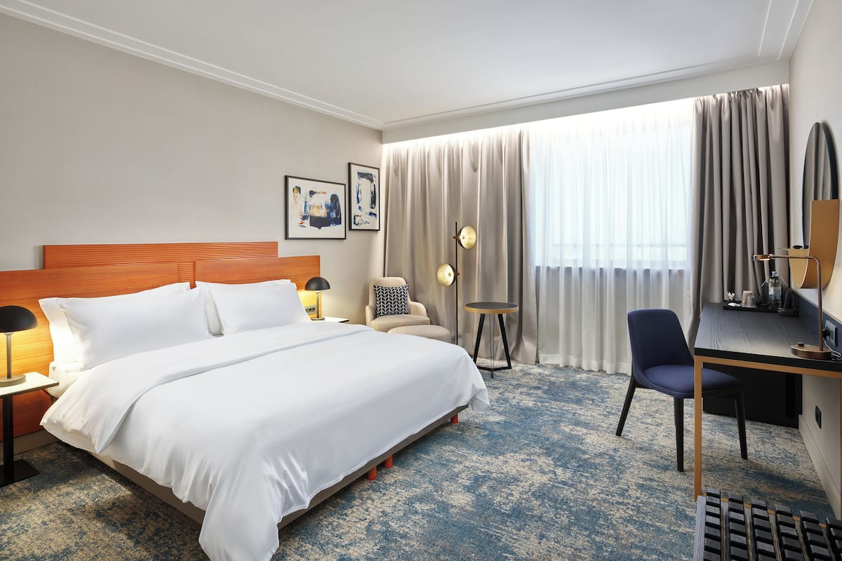 A hotel room with a large bed, a desk with a chair, a small table, and a window with curtains.