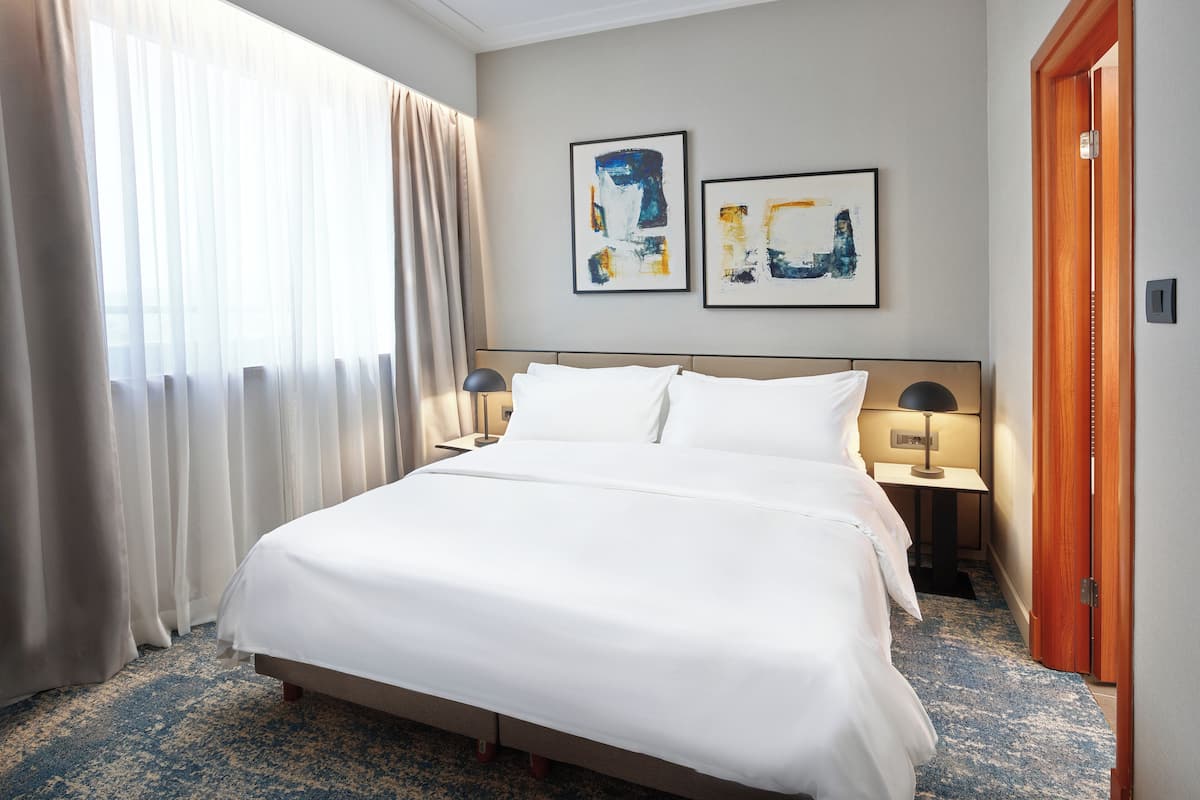 A hotel room with a large bed, two bedside tables with lamps, a wooden door, and two abstract paintings on the wall.