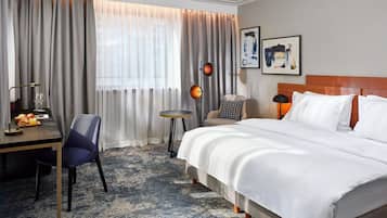 A hotel room with a bed, a desk with a chair, a small table, and a lamp.
