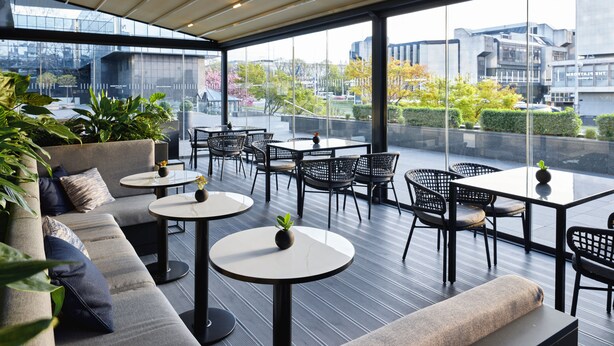 Un espacio moderno al aire libre con sofás, mesas redondas y sillas, y vistas a la ciudad a través de amplios ventanales.