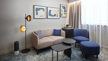 Una habitación de hotel moderna con un sofá color beige, un sillón azul, una mesita redonda pequeña, una lámpara de pie y arte abstracto en la pared.