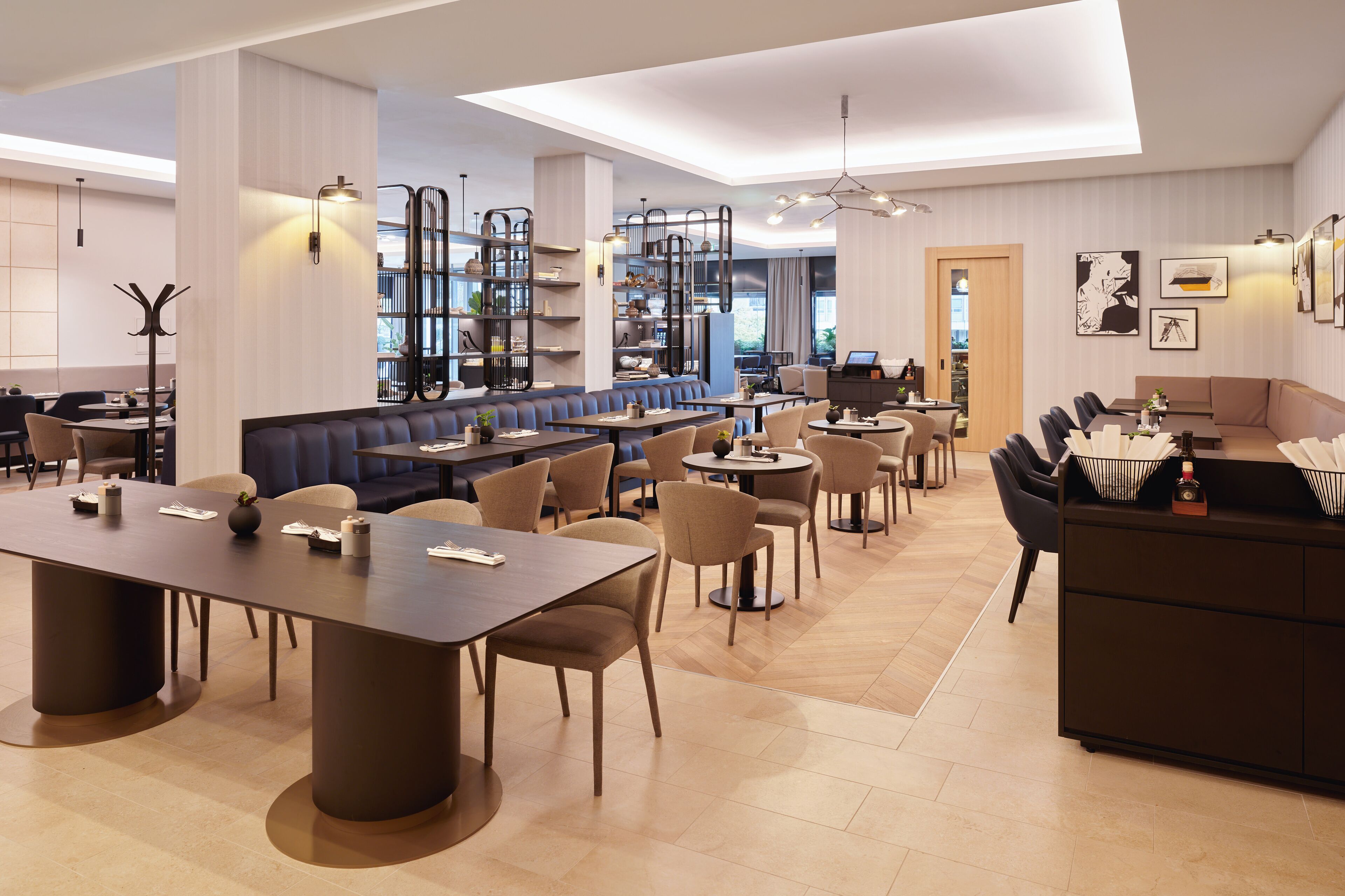 A modern restaurant with a long communal table, several smaller tables, and a bar area.