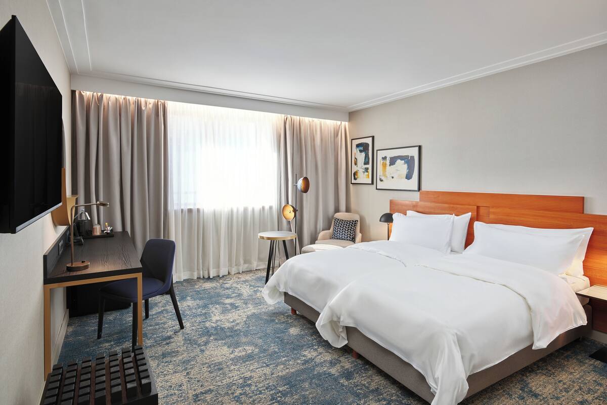 A hotel room with a bed, desk, chair, and a large window with curtains.
