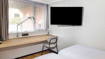 Habitación de hotel con cama, escritorio con silla, televisor montado en la pared y ventana con cortinas.
