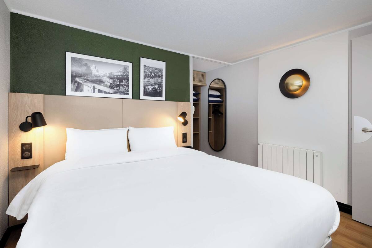 A neatly made bed with white linens, a headboard with a wooden finish, two wall-mounted lamps, and two framed black-and-white photographs above the bed.
