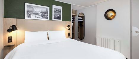 A neatly made bed with white linens, a headboard with a wooden finish, two wall-mounted lamps, and two framed black-and-white photographs above the bed.