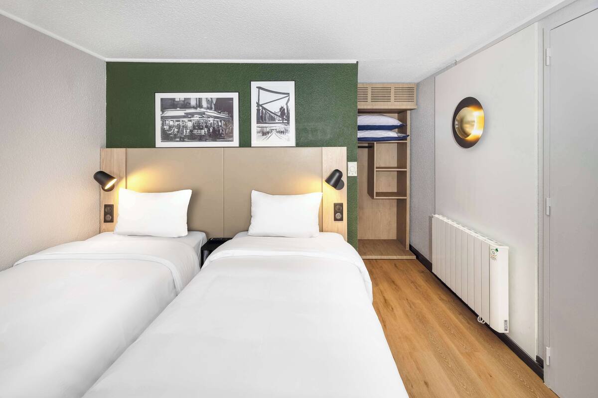A hotel room with two single beds, a green accent wall, a radiator, and a small shelving unit with folded towels.