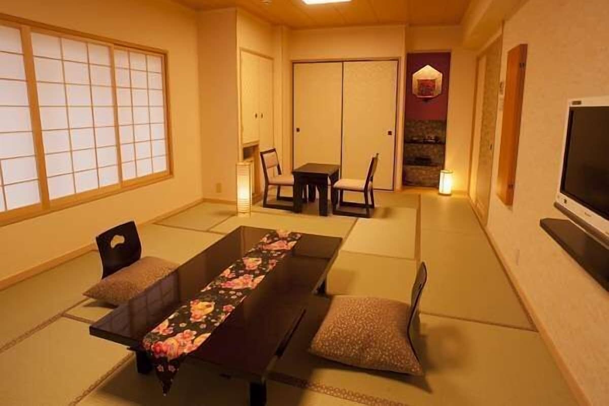 A traditional Japanese room with tatami flooring, a low table with a floral table runner, two chairs, a TV on the wall, and a sliding door.