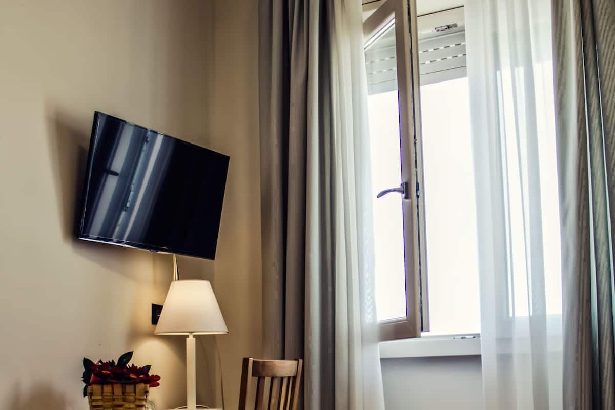 Una camera d'albergo con un letto, una scrivania con una lampada, una sedia, una televisione a muro e una finestra con tende.
