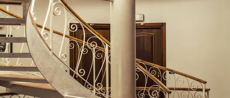 Uma escadaria em espiral com grades de metal ornamentadas e degraus de madeira, que leva a um andar superior com vista do céu através de grandes janelas.