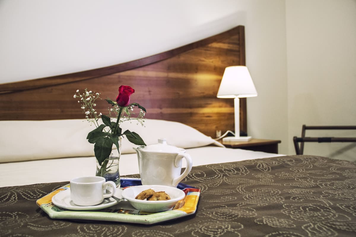 Un letto rifatto con cura, una testiera in legno, una lampada da comodino e un vassoio con una tazza, un piccolo vaso con una rosa e un teiera.