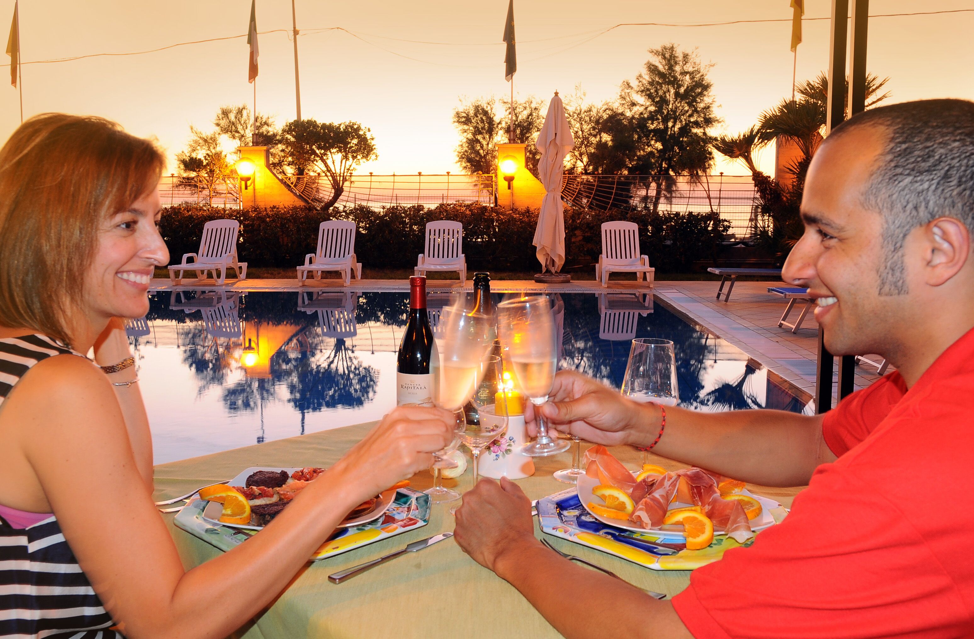 Une table à manger avec des assiettes remplies de nourriture et des boissons, une piscine en arrière-plan et des chaises d’extérieur.