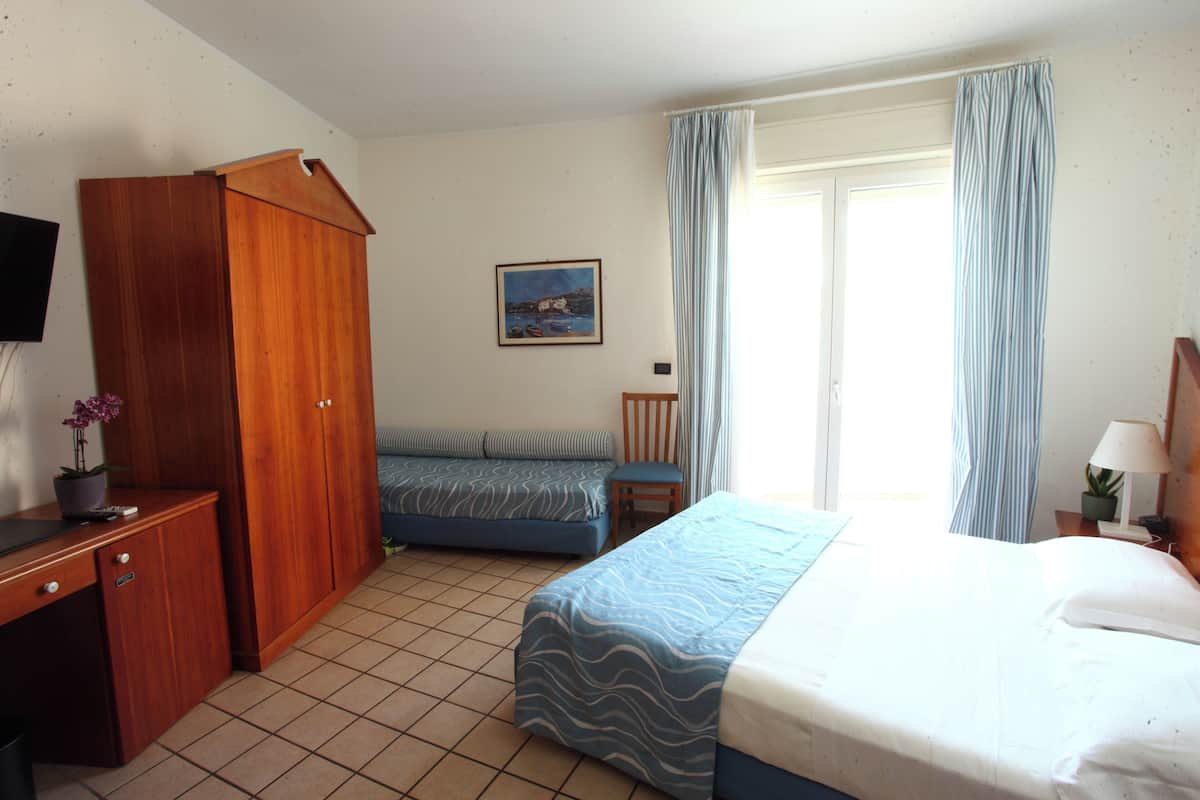 Una camera d'albergo con un letto, una scrivania, un armadio, una sedia, una televisione e una finestra con tende.