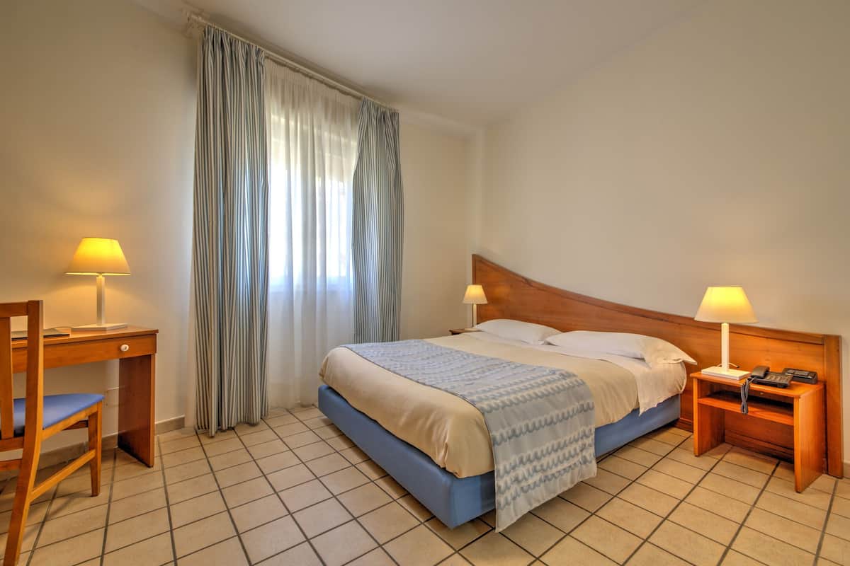 Una camera d'albergo con un letto, due comodini con lampade, una scrivania e una sedia.