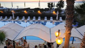 Zona de comedor junto a la piscina con mesas y sillas bajo sombrillas, con vistas a la piscina y al mar.