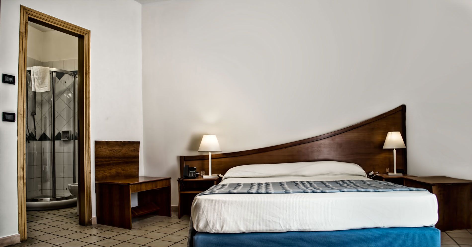 Habitación de hotel con cama, dos mesitas de noche, cabecero de madera, lámpara y espejo.