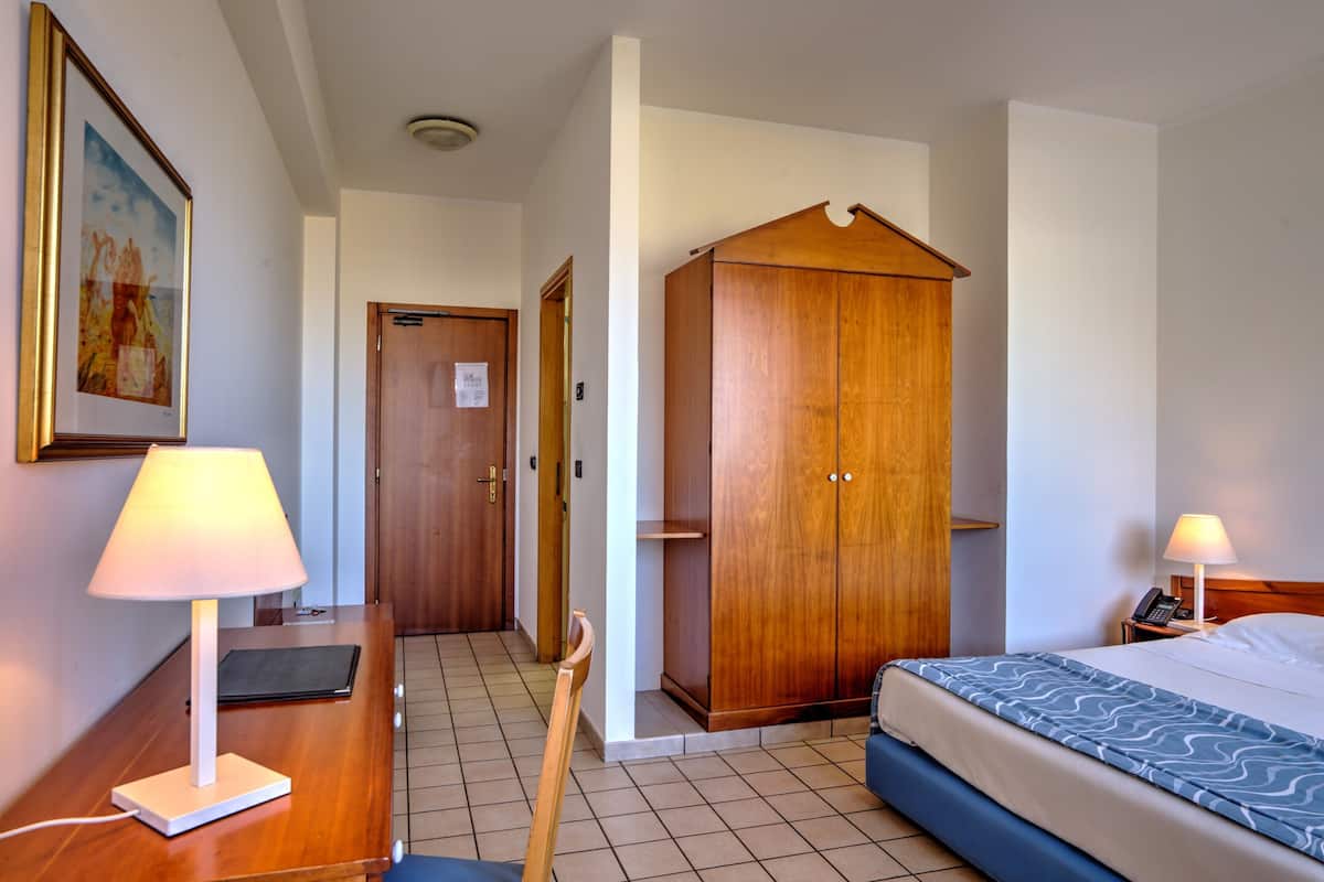 Una camera d'albergo con un letto, un comodino, una lampada, un armadio in legno e una porta che dà su un'altra stanza.