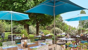 An outdoor dining area with tables and chairs, blue umbrellas, and a garden setting. - Kyriad Direct Le Bourget - Gonesse (Gonesse)