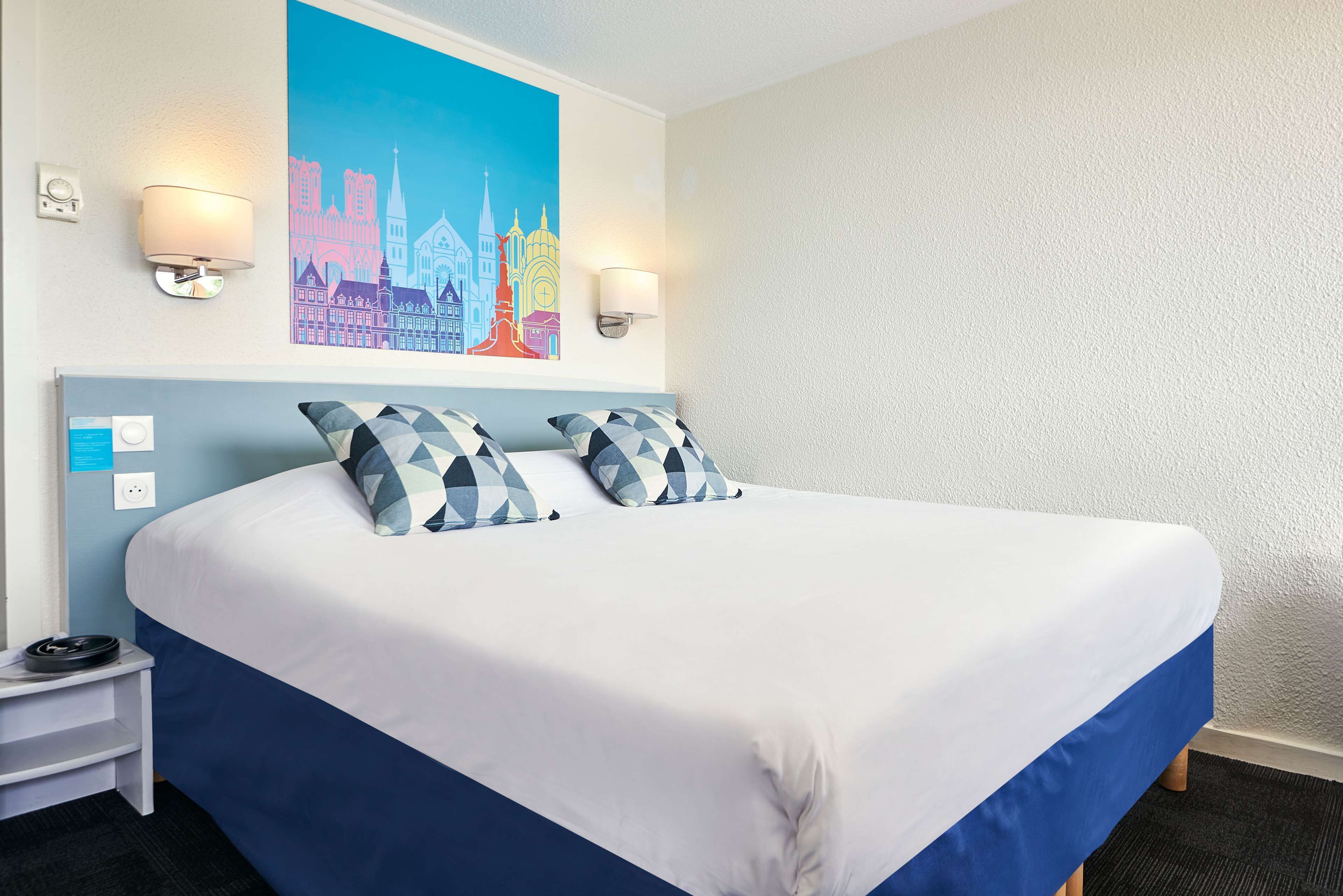 Quarto de hotel com uma cama grande, duas luminárias fixadas na parede, uma pintura de paisagem urbana e uma pequena mesa de cabeceira com um telefone.
