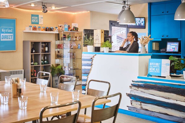 L’intérieur d’un restaurant, avec une table à manger en bois, des chaises, un comptoir derrière lequel se trouve une personne, des étagères remplies de bouteilles et un mur bleu orné d’une enseigne.
