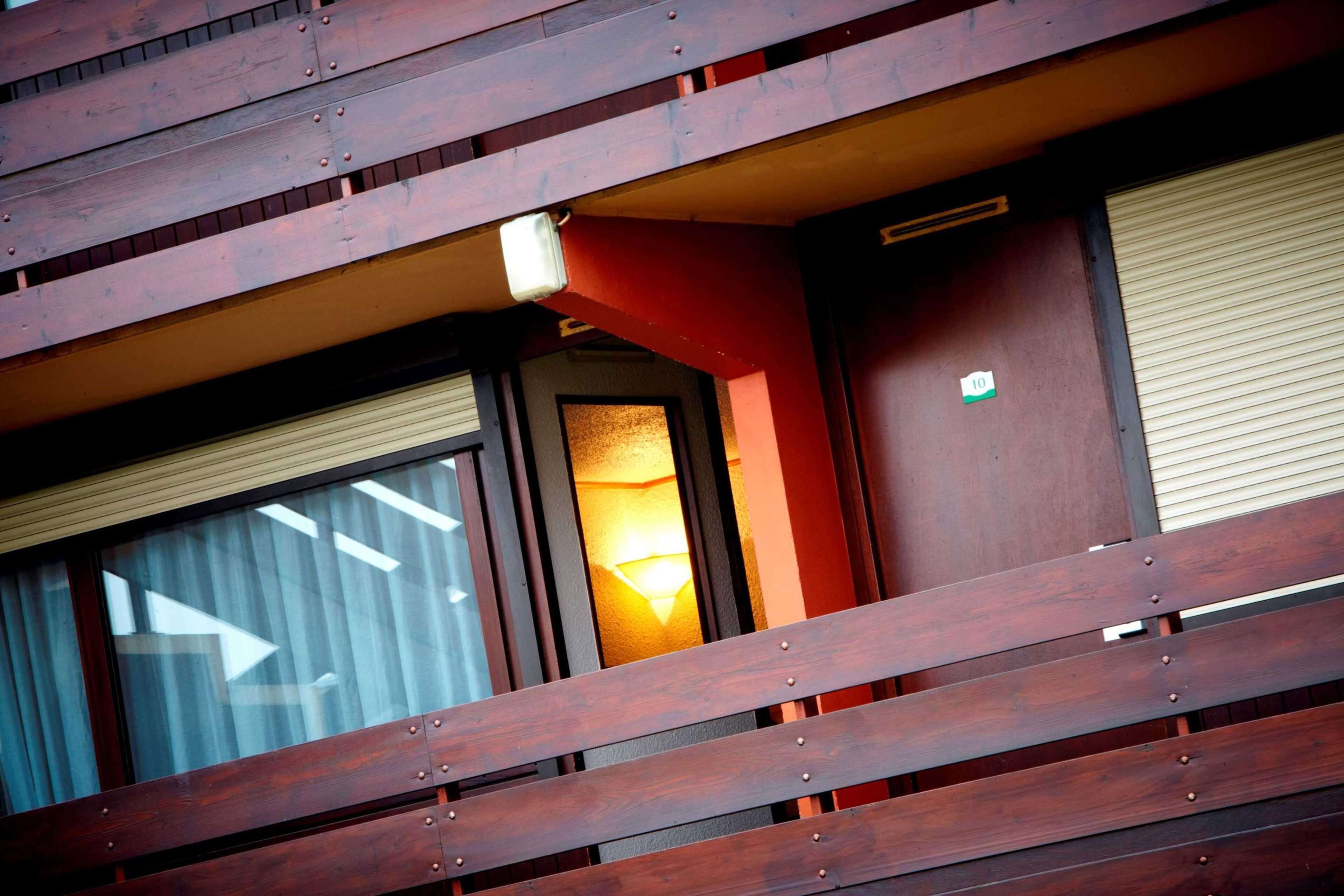 Un balcón de madera con vistas al interior de un edificio, incluyendo una ventana con persianas y una puerta con un cartel.