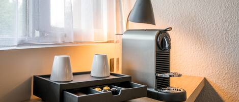 Una máquina de café con un portavasos, un cajón para cápsulas de café y una lámpara sobre una mesa de madera cerca de una ventana.