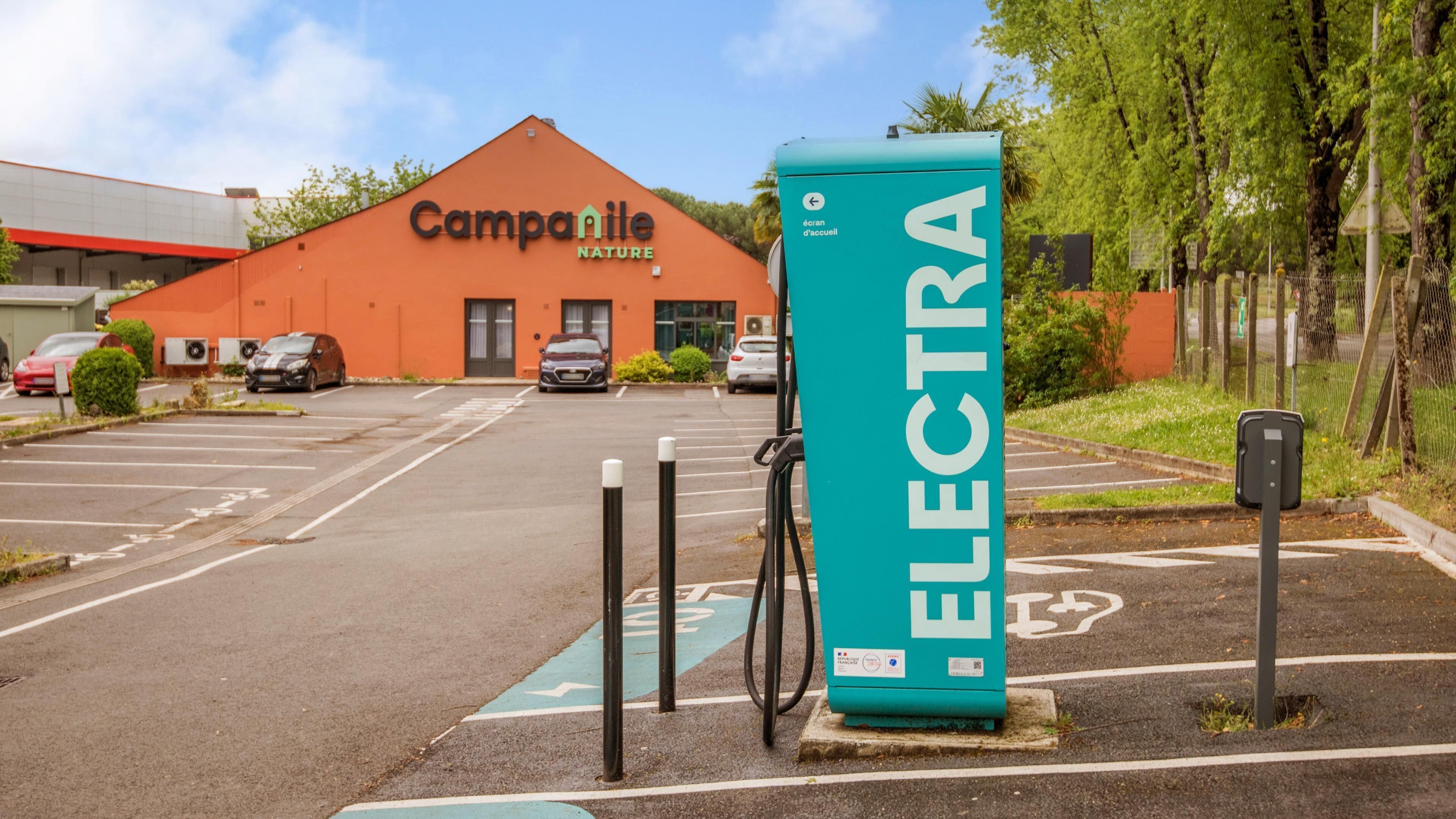 A parking lot with a green electric vehicle charging station, a building labeled 'Campanile Nature,' and a clear sky.