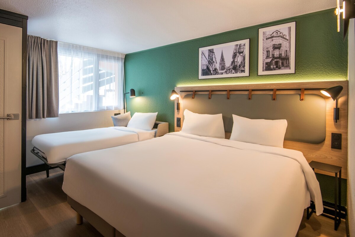 Habitación de hotel con dos camas, una ventana con cortinas, una pared verde y dos cuadros enmarcados.