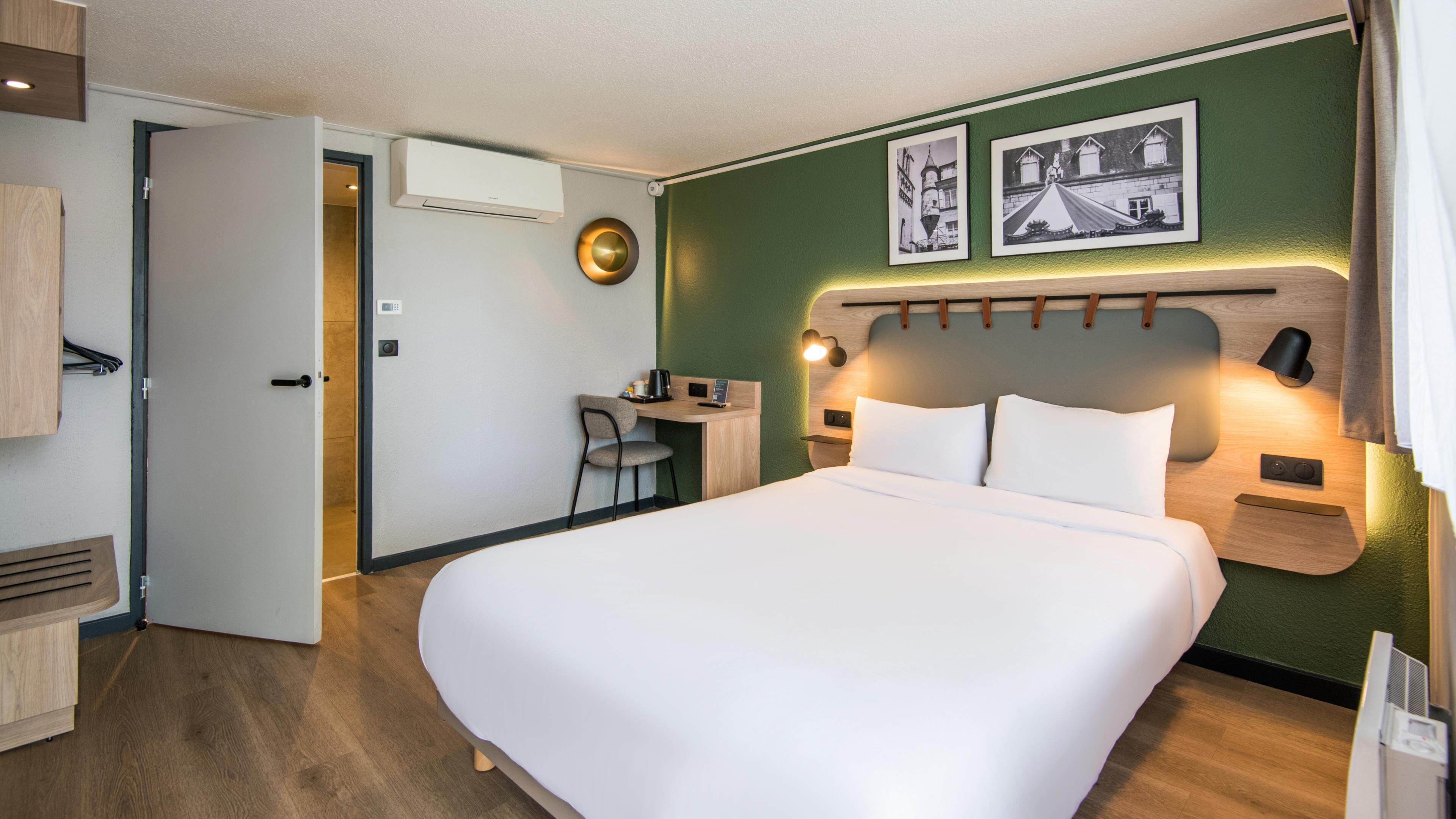 A hotel room with a bed, desk, chair, and a wall-mounted air conditioner.