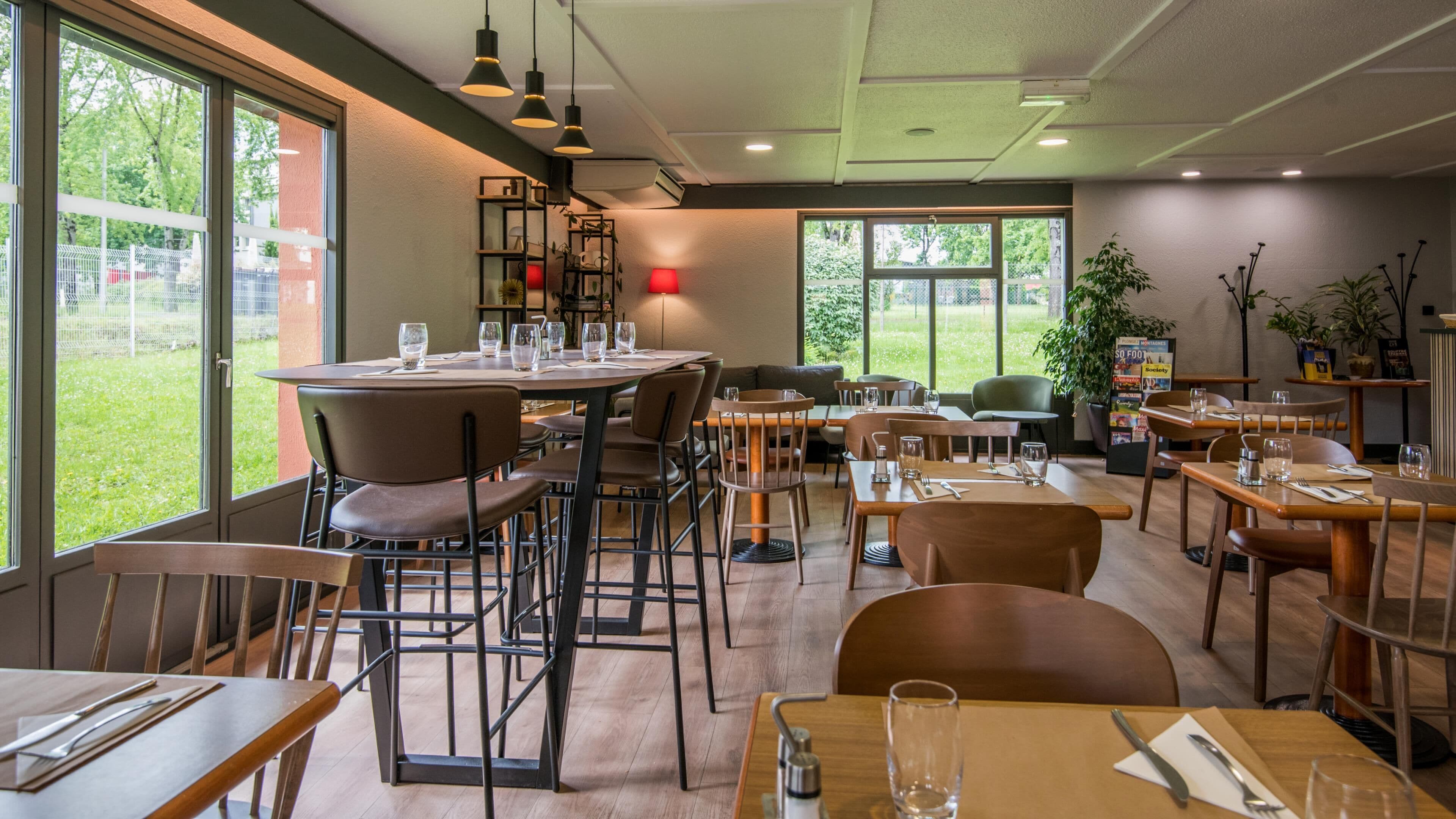 A restaurant with multiple tables and chairs, large windows, and indoor plants.