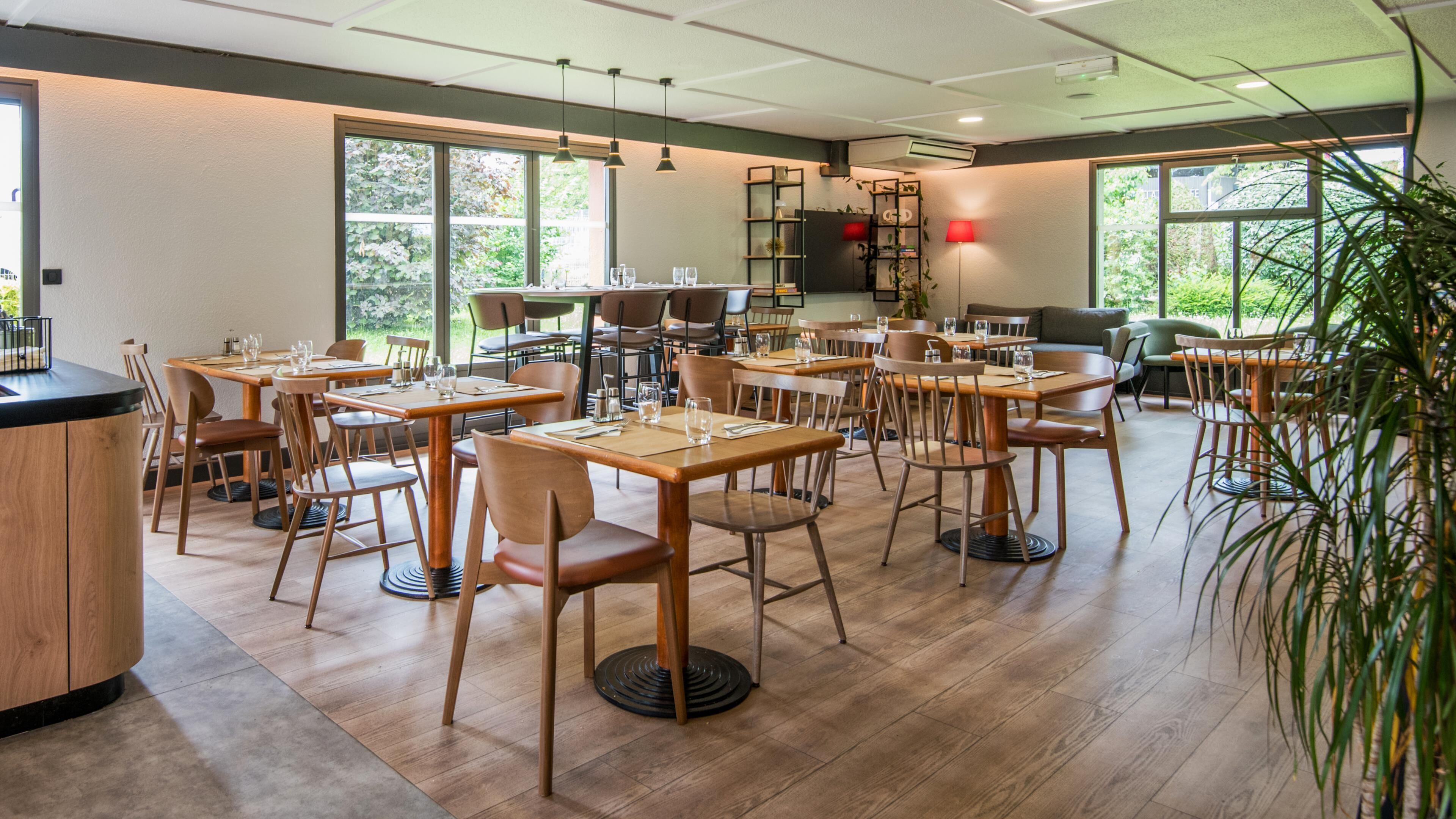 A modern dining area with multiple tables and chairs, large windows, and a kitchen area visible in the background.