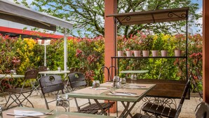 Un patio al aire libre con mesas y sillas, una pérgola con detalles decorativos y abundante follaje al fondo.