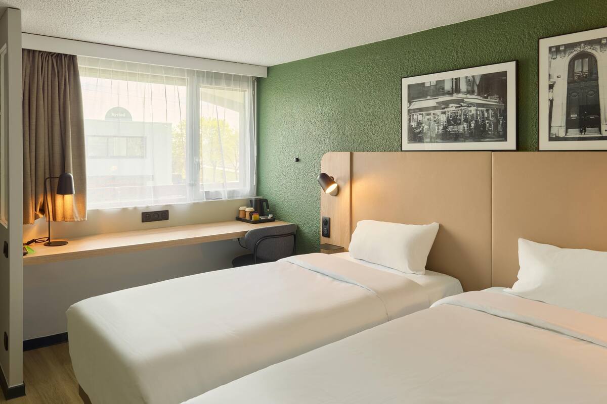 Habitación de hotel con dos camas, un escritorio con lámpara, una ventana con cortinas y una pared verde con textura.