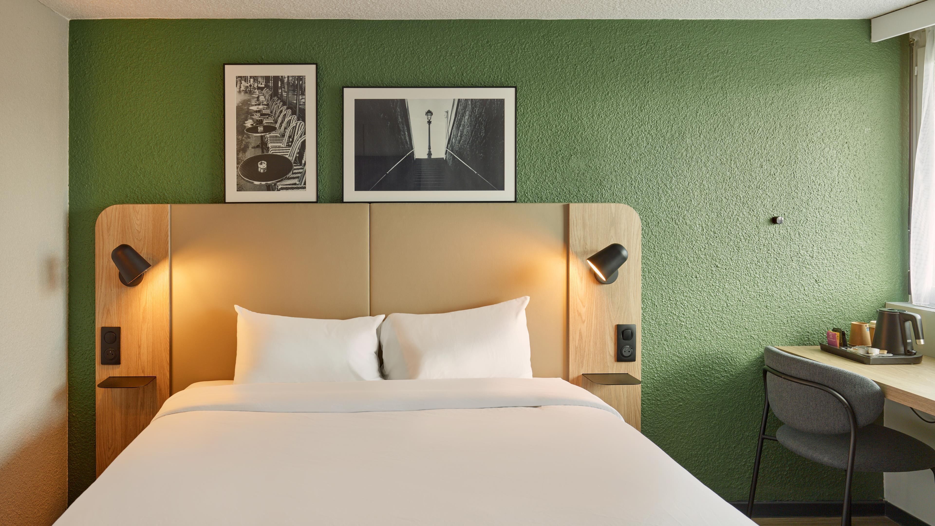 Quarto de hotel com uma cama, uma escrivaninha e duas imagens emolduradas na parede.