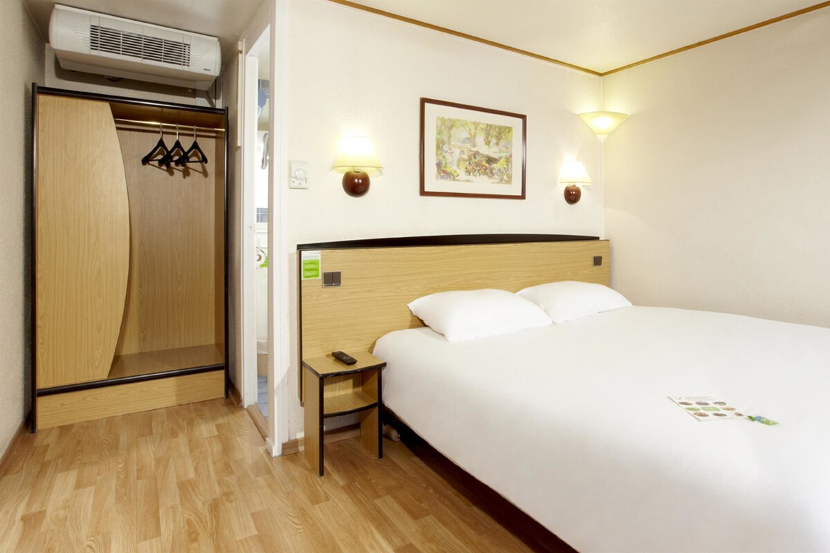 A hotel room with a wooden wardrobe, a bedside table with a remote control, a bed with white bedding, and two wall-mounted lamps.