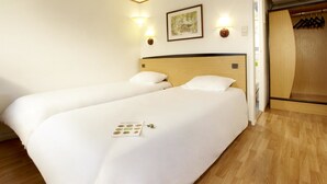 A hotel room with two beds, a wooden headboard, wall-mounted lights, a framed picture, and a wooden closet with hangers.