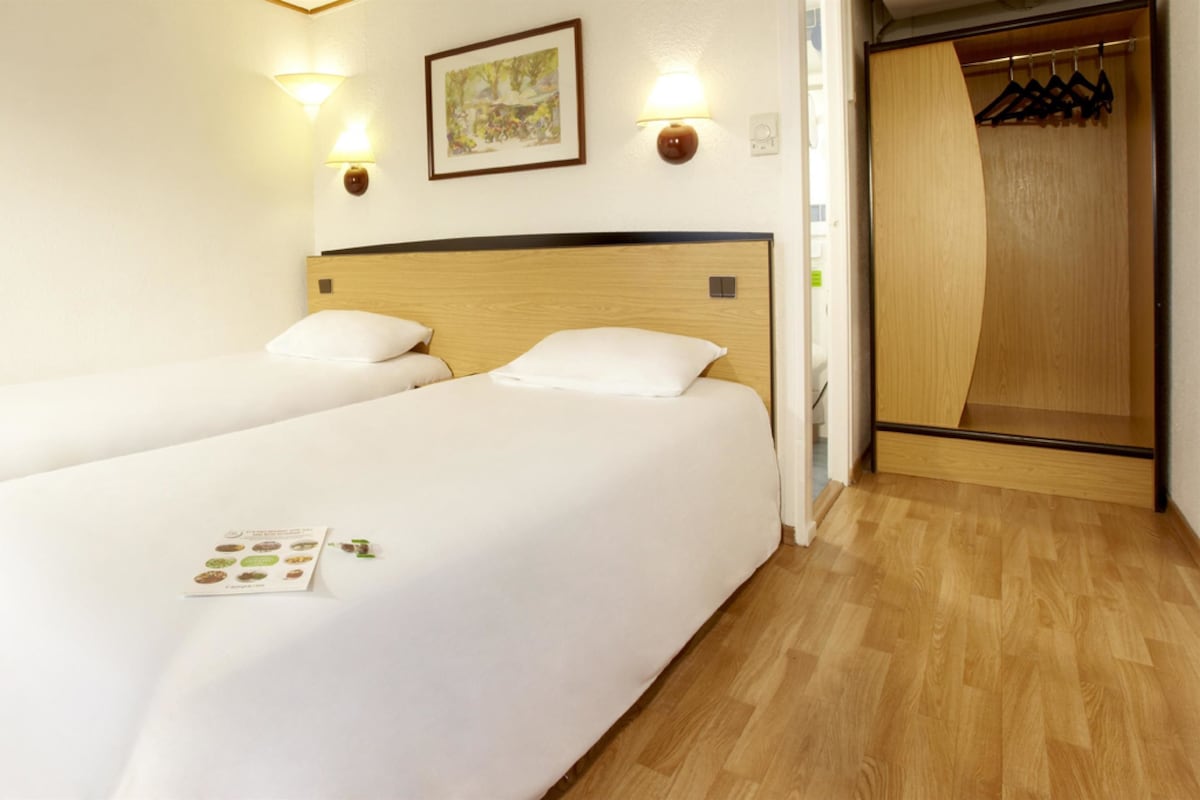 A hotel room with two beds, a wooden headboard, wall-mounted lights, a framed picture, and a wooden closet with hangers.