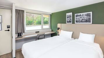 A hotel room with a bed, a desk, a chair, a window with curtains, and two black and white photographs on the wall.