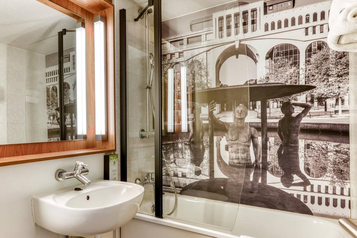 Un baño con un espejo grande, un lavabo, cabina de ducha con puertas de cristal y vistas a un edificio a través del cristal de la ducha.