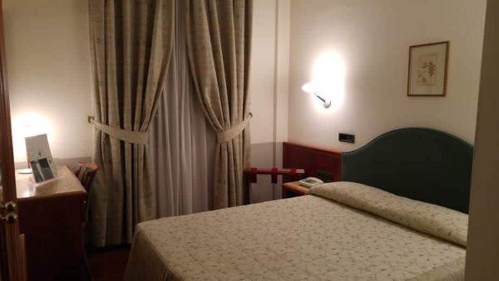 Una habitación de hotel con cama, mesita de noche, lámpara, cortina y aplique de pared.