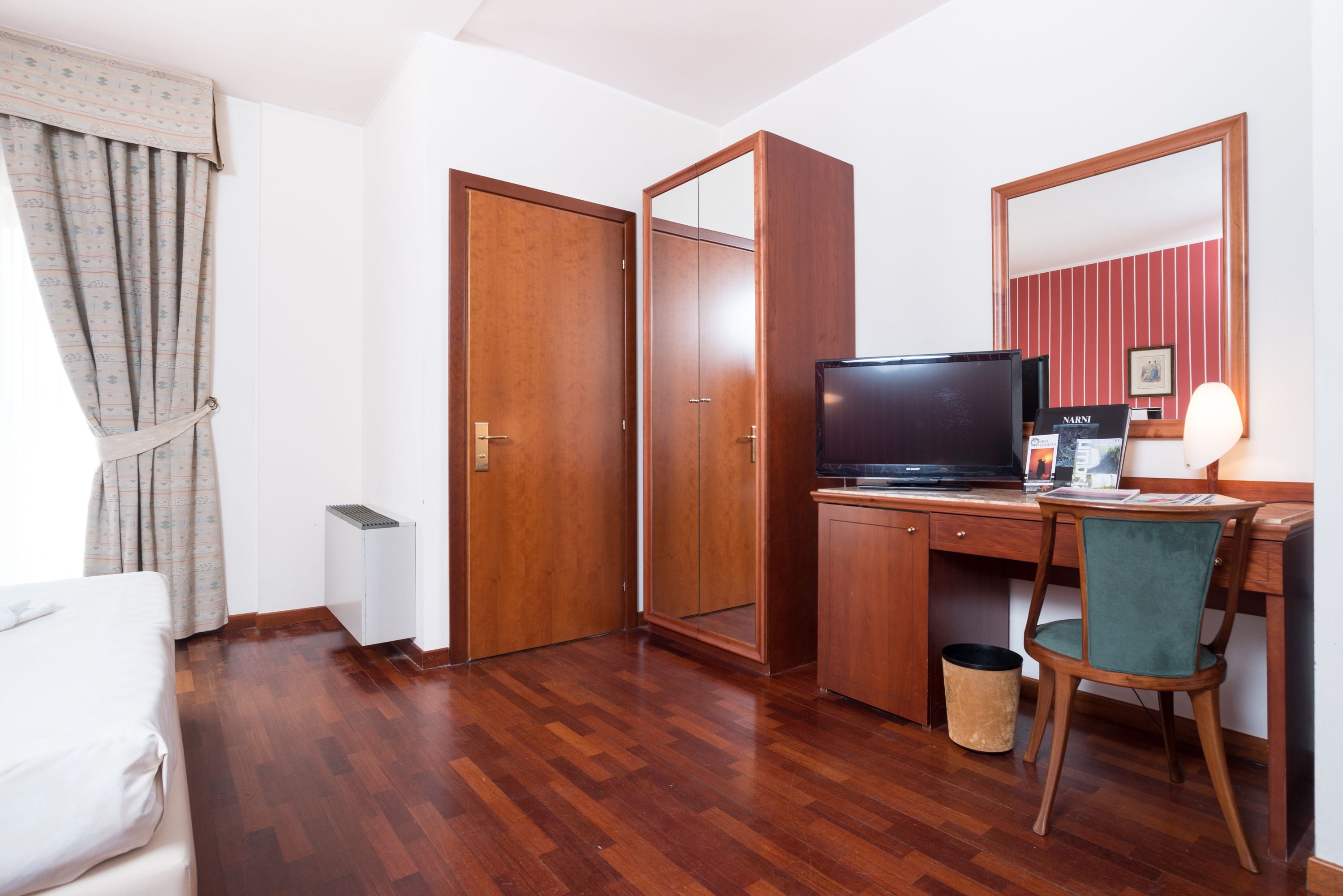 Una habitación de hotel con suelo de madera, un escritorio con computadora, televisor y espejo.