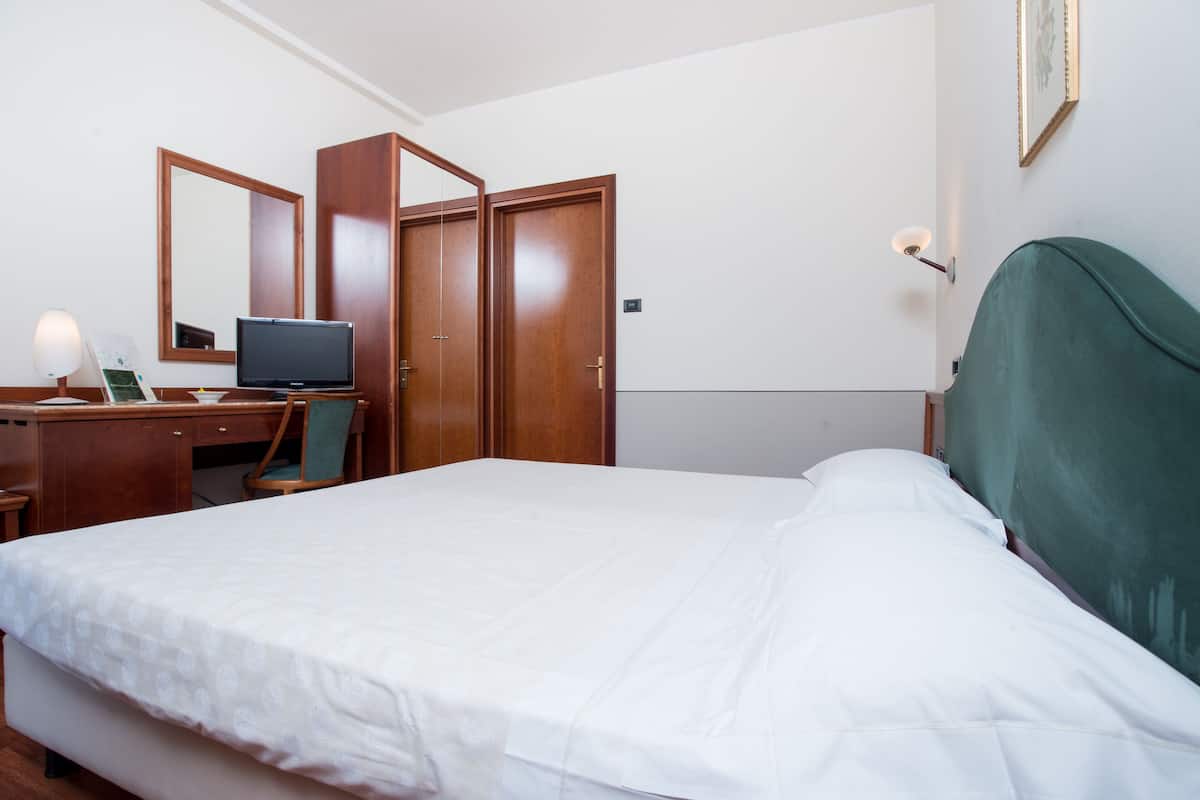 Hotellihuone, jossa on sänky, työpöytä tietokoneella, tuoli, vaatekaappi ja yöpöytälamppu.