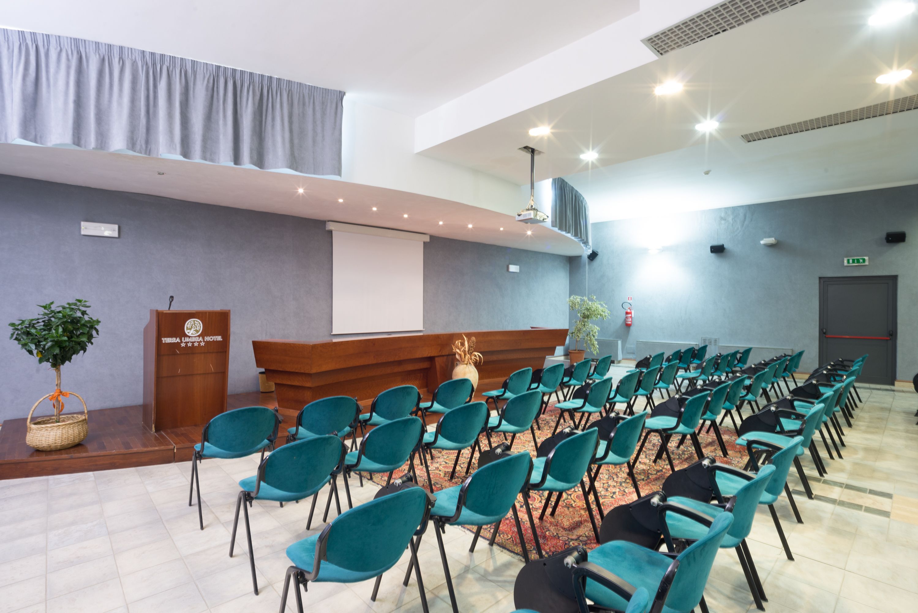 Ein Konferenzraum mit Reihenweise angeordneten türkisfarbenen Stühlen, einem Rednerpult und einer Projektionsleinwand.